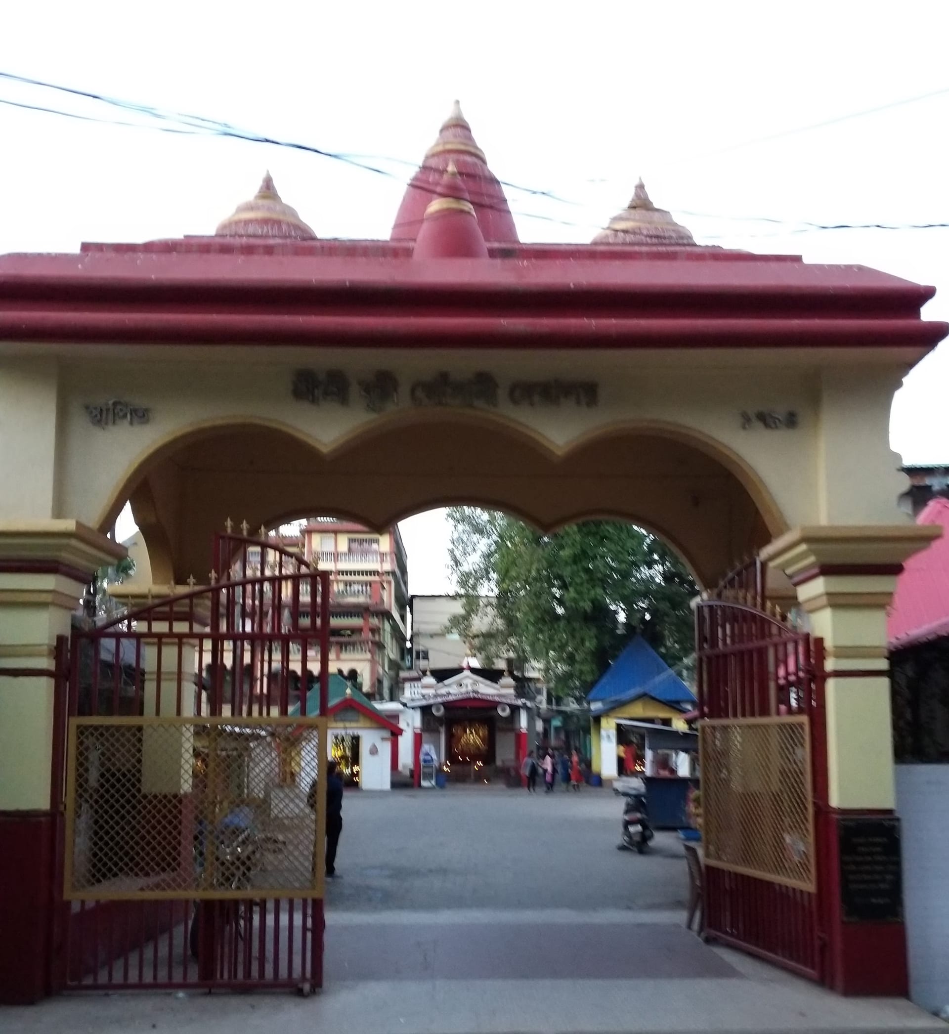 The cream-and-red ornamental gateway of the Burhi Gosani Devalaya, three red temple spires rising behind it, the courtyard shrines seen through the open gate