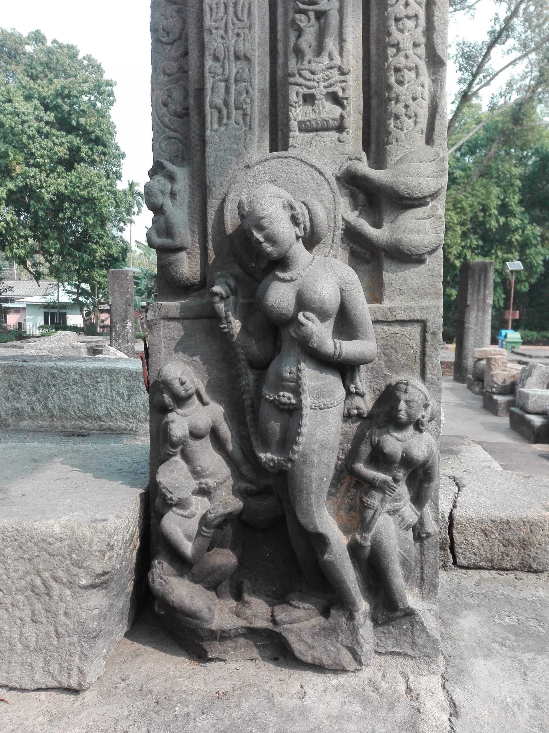 A slender, gently swaying river-goddess with a halo, holding a garland and standing on her mount at the foot of a doorframe jamb, flanked by two smaller attendant figures, with bands of foliate carving rising above.