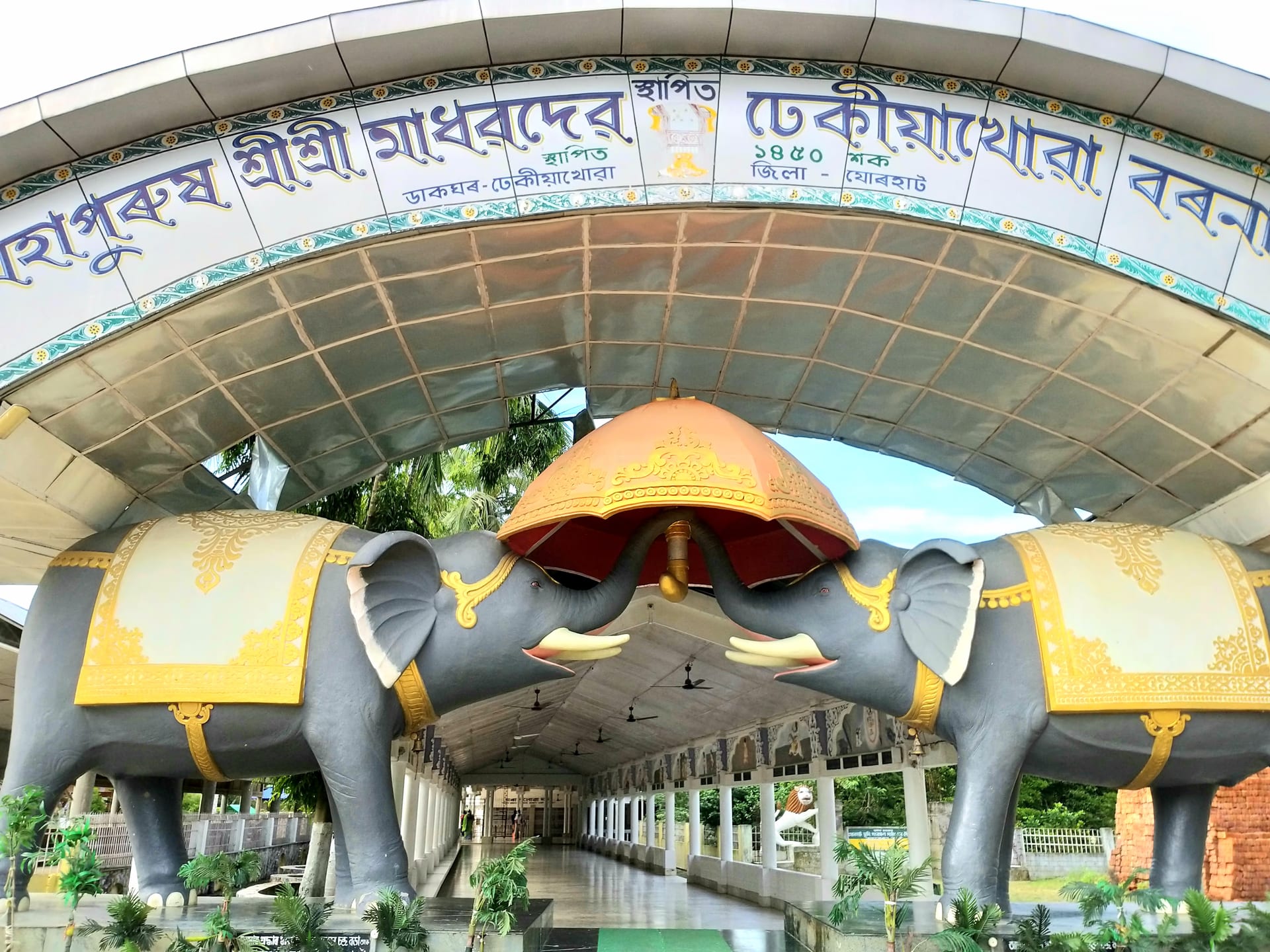 Ornate gateway of Dhekiakhowa Bornamghar flanked by two large elephant sculptures, with a covered walkway leading to the prayer-hall behind
