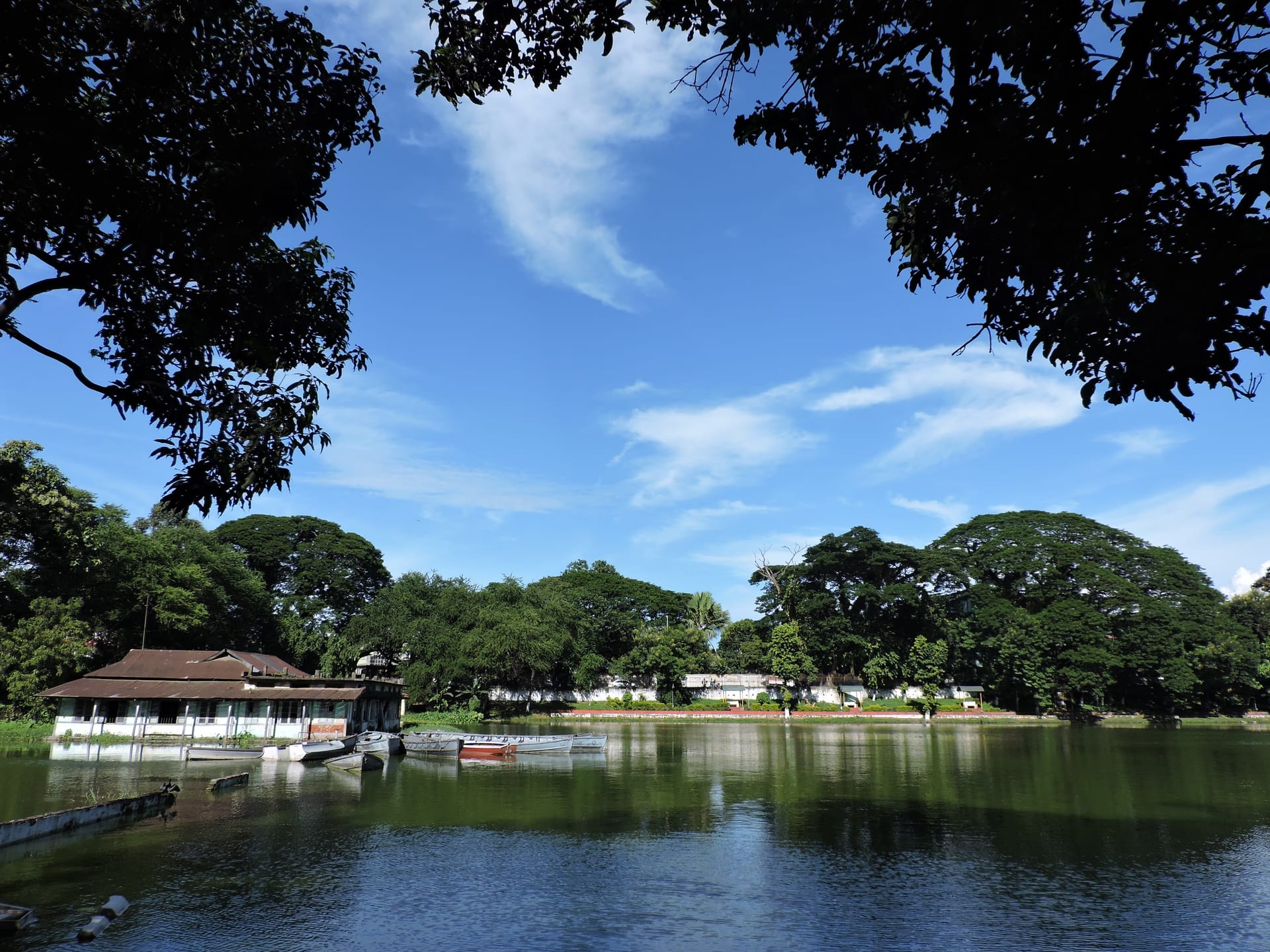 The long tank of Dighalipukhuri in Guwahati, wooden boats moored by an old boathouse on the left and the tree-lined far bank under a blue sky, still green water reflecting the trees