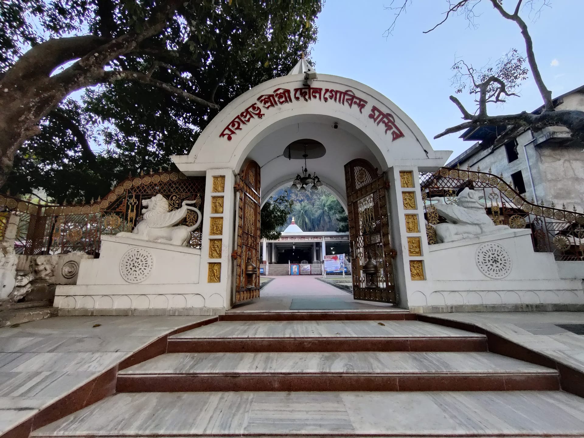 The arched entrance gateway to Doul Govinda temple: a whitewashed arch carrying the temple's name in red Assamese lettering, flanked by carved white lion sculptures and golden filigree gates, with marble steps leading up and the white-domed Govinda shrine framed in the opening beyond