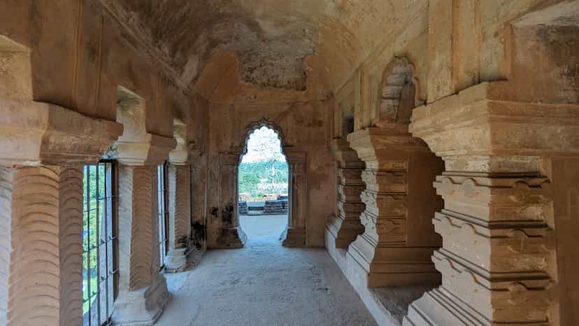 A vaulted upper gallery inside the Kareng Ghar palace at Garhgaon: a row of fluted masonry columns with window grilles runs down the left, carved pilasters down the right, and a scalloped arch doorway at the far end opens onto daylight