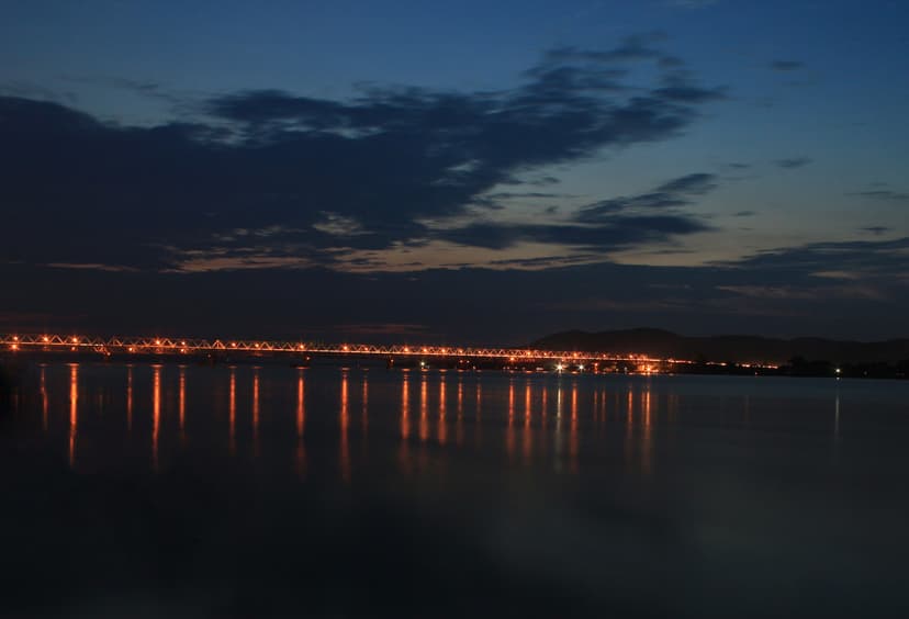 The Saraighat rail-and-road bridge stretched across the Brahmaputra at blue hour, its long line of orange lights mirrored in the still dark water beneath a deep-blue sky streaked with cloud, low hills on the far bank