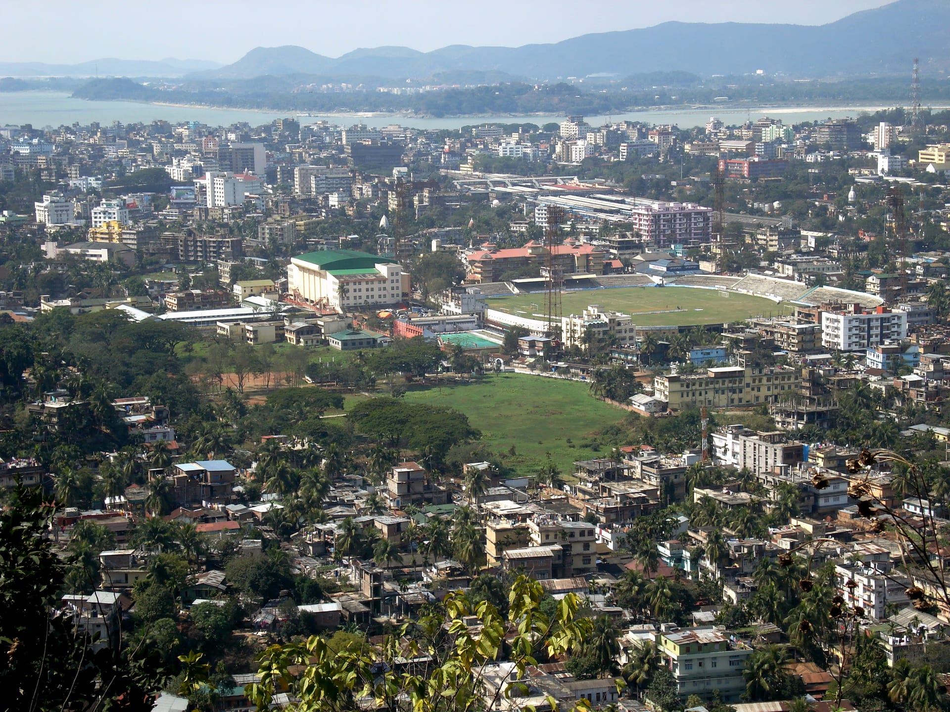 Guwahati spread below a hilltop viewpoint: dense low buildings and palms, the green oval of the Nehru Stadium at centre, the wide Brahmaputra and blue hills on the horizon