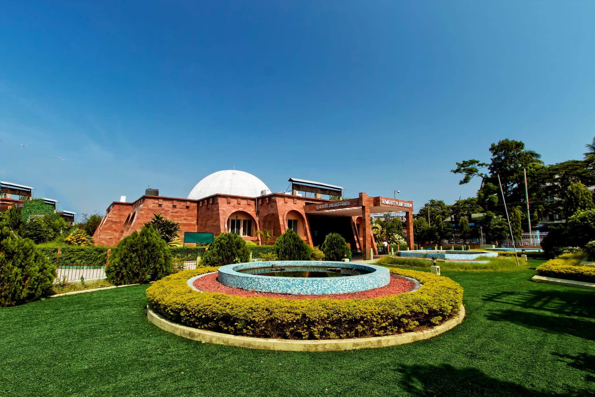 The white dome and sloped red-brick wings of the Guwahati Planetarium under a deep blue sky, lawns and a round mosaic fountain in front