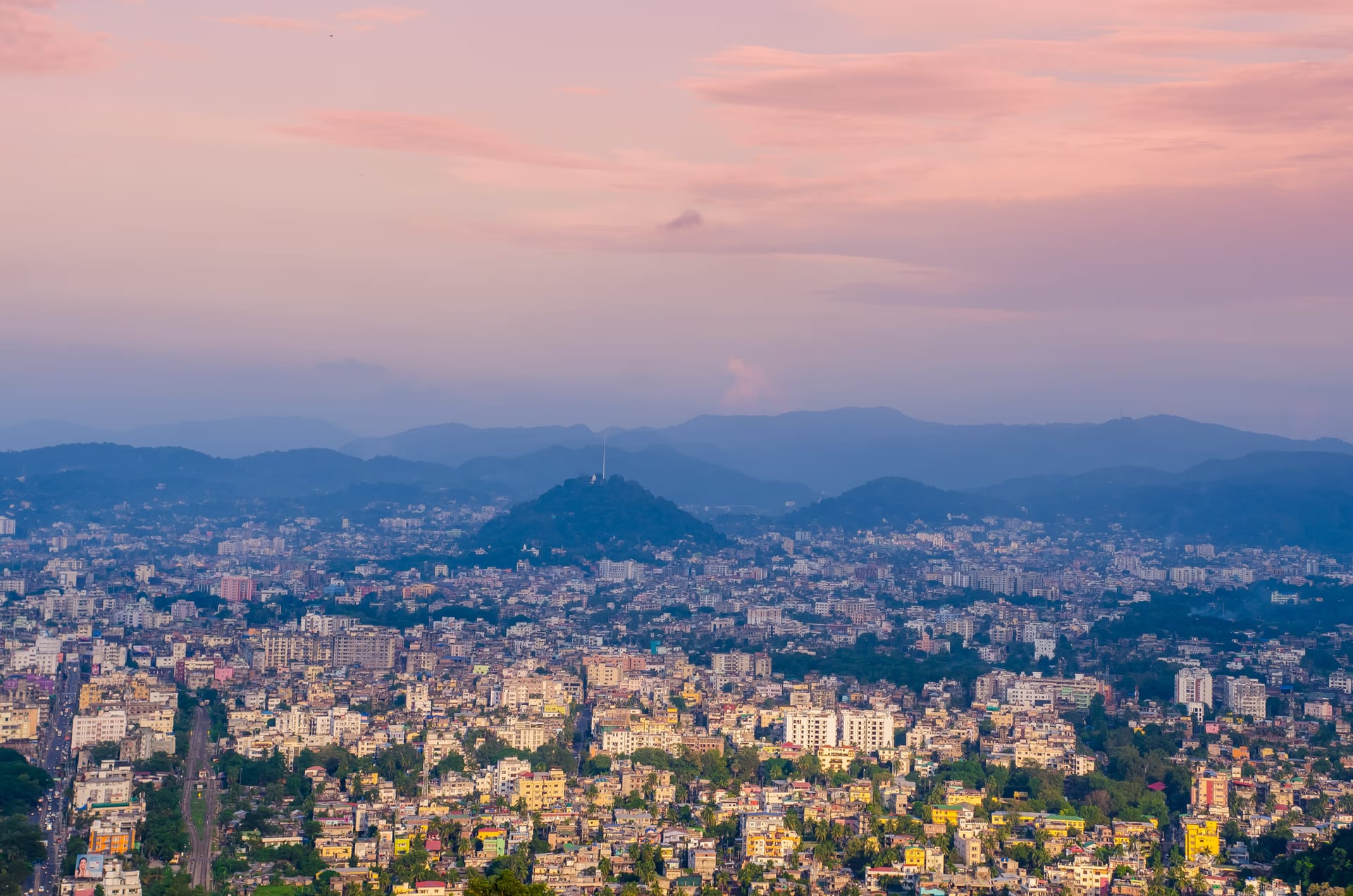 Guwahati at dusk seen from high on the Nilachal hill: a sea of buildings around a lone wooded hillock, ranges of blue hills behind, under a pink evening sky
