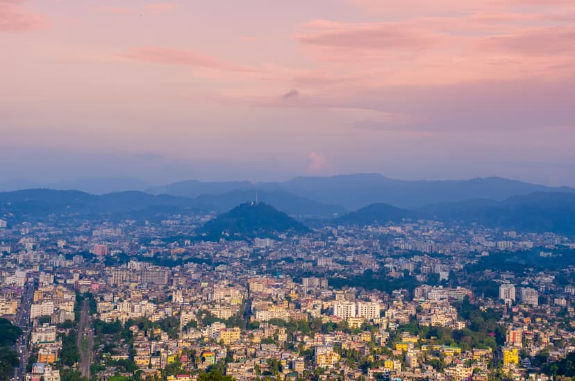 Guwahati at dusk seen from high on the Nilachal hill: a sea of buildings around a lone wooded hillock, ranges of blue hills behind, under a pink evening sky