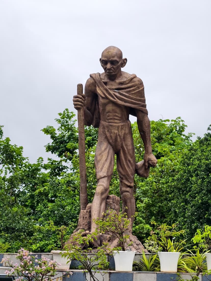 A weathered bronze-toned statue of Mahatma Gandhi mid-stride with staff, against trees and an overcast sky at Gandhi Mandap