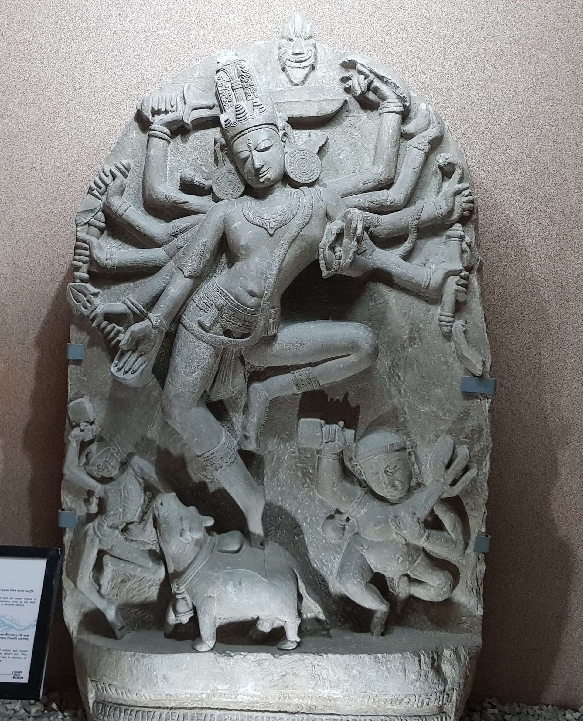 A tall grey stone stele carved with a many-armed dancing Shiva, knee raised across his body, ringed by his own arms holding a trident, drum and other attributes, with attendant figures and the bull Nandi below his feet