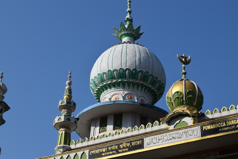 The white ribbed dome of the Powa Mecca dargah at Hajo, ringed with green lotus-petal moulding and topped by an ornamental finial, beside a gold crescent-topped dome and slender minarets against a clear blue sky
