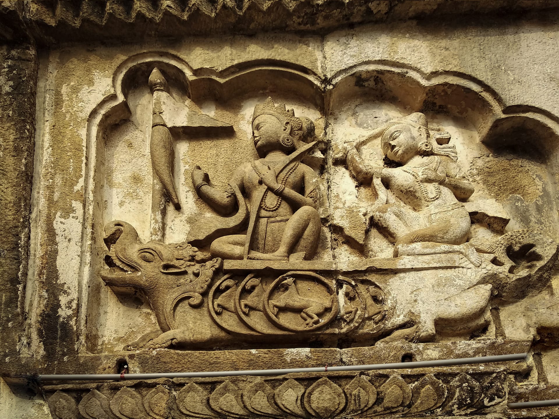 A sandstone relief panel set in a cusped niche on the temple wall: a crowned figure riding a makara, a horned aquatic beast, and holding a stringed instrument, faced by a second seated figure, above a band of carved lotus-petal ornament