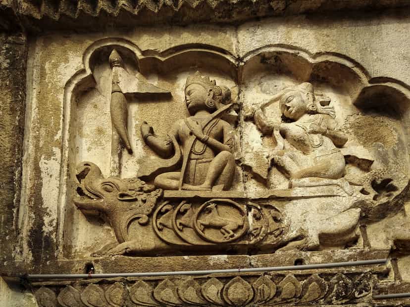 A sandstone relief panel set in a cusped niche on the temple wall: a crowned figure riding a makara, a horned aquatic beast, and holding a stringed instrument, faced by a second seated figure, above a band of carved lotus-petal ornament