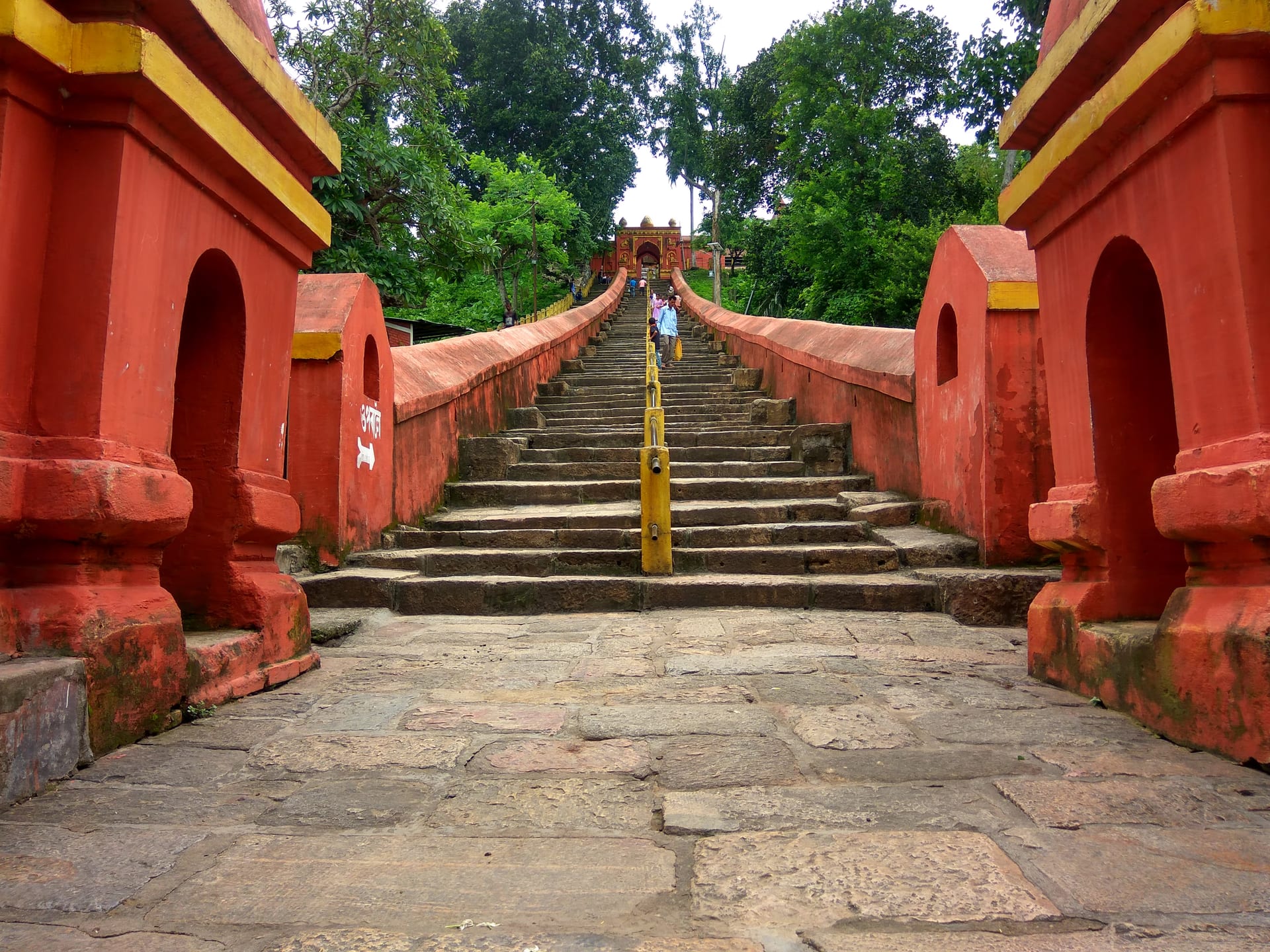 Steep flight of grey stone stairs climbing between red-and-yellow painted gate pillars toward the Hayagriva Madhava temple on the Manikut hill, a yellow railing down the middle