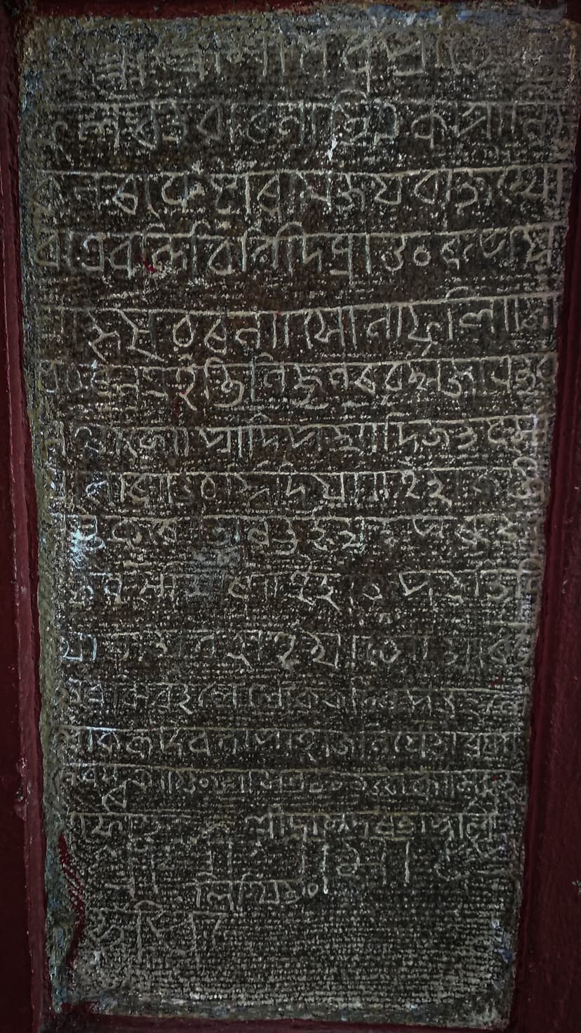 Tall weathered dark stone slab set in a red wall, densely carved with many lines of Assamese-Bengali script beginning with Shri Shri