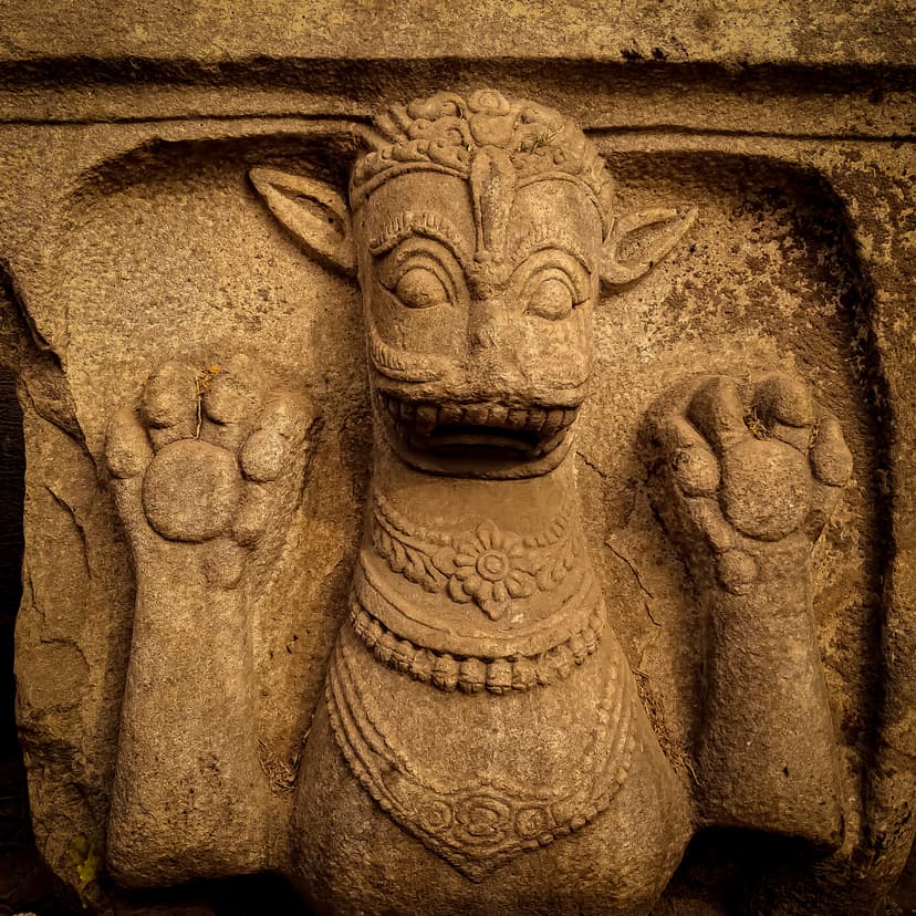 Close-up of a weathered stone relief of a fierce animal-faced guardian with raised paws, jeweled necklaces and crown, set in a niche