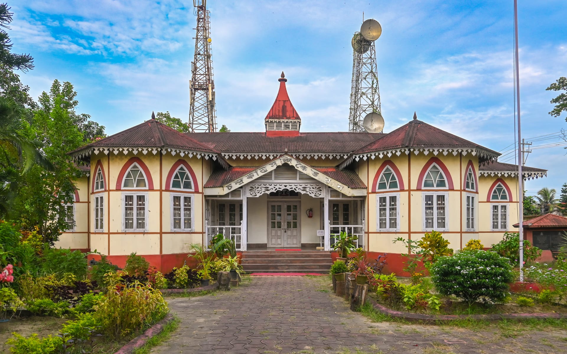 An ornate single-storey heritage building with a steep tiled gable roof, a central red-domed turret, arched windows and a symmetrical verandah, fronted by a paved path and garden under a blue cloud-streaked sky