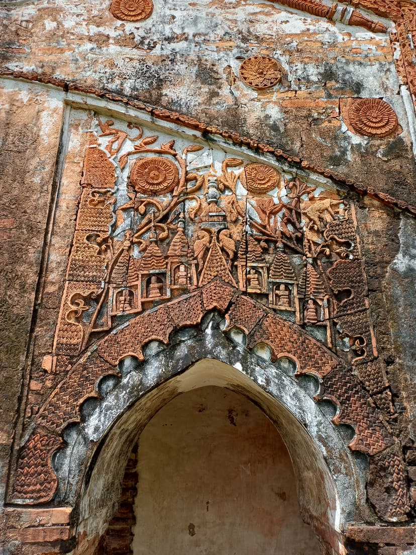 Terracotta relief above a cusped arch on the Ghanashyam Dol at Joysagar: miniature temple gables, birds, climbing monkeys, and floral medallions in weathered clay