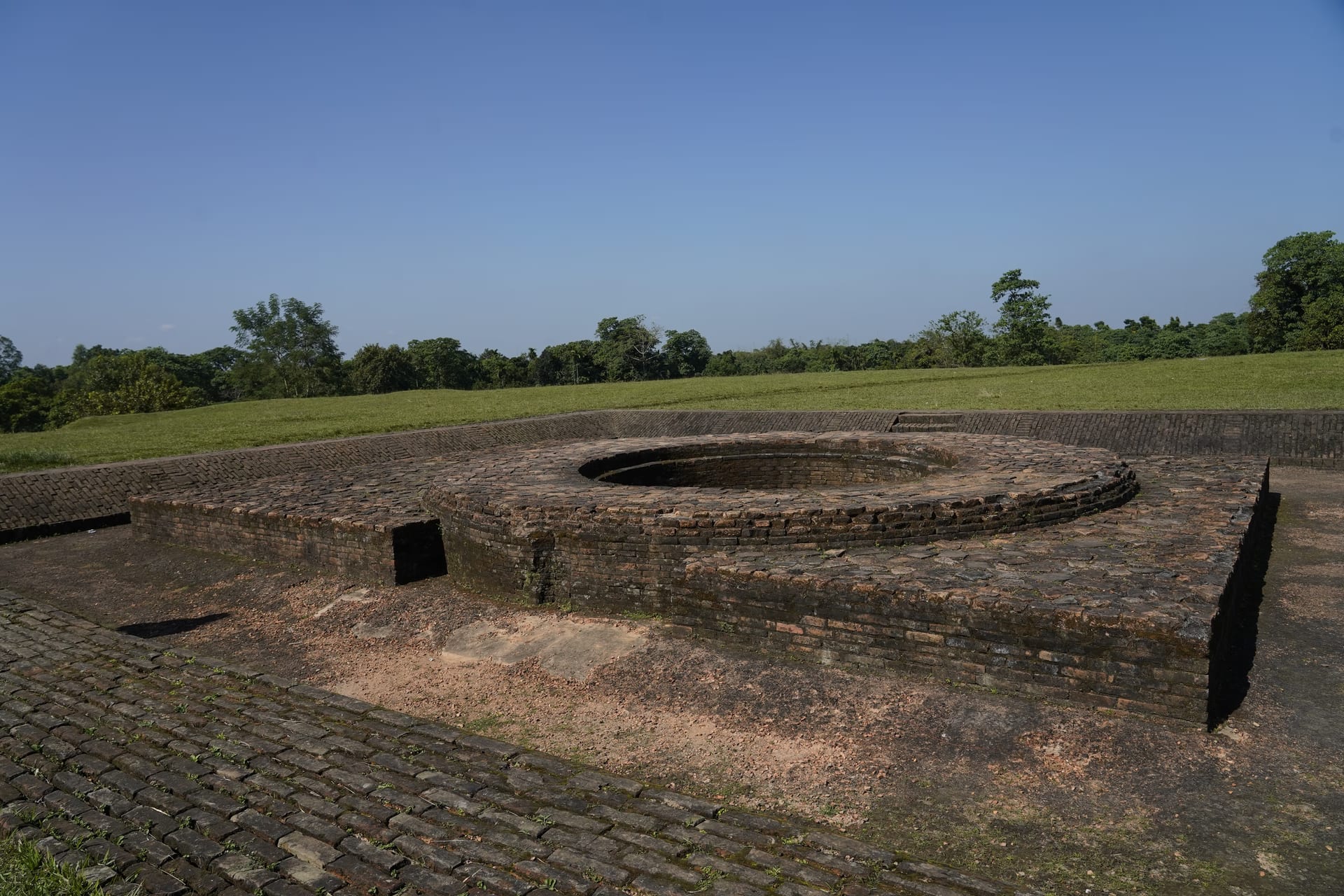 A tiered ring of weathered red-brick courses around a sunken circular pit on a brick-paved platform at the Rajpat mound, set into a green grassed earthwork under a clear blue sky with a tree line behind