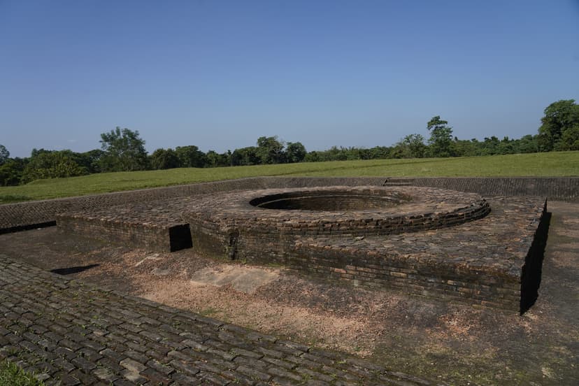 A tiered ring of weathered red-brick courses around a sunken circular pit on a brick-paved platform at the Rajpat mound, set into a green grassed earthwork under a clear blue sky with a tree line behind