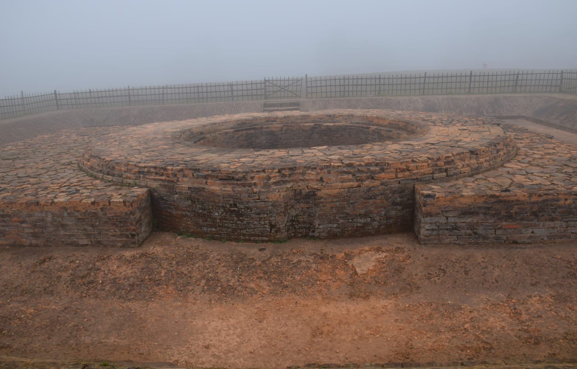 A low, tiered ring of warm red brick rising from a brick-paved mound, dense morning mist erasing the fields and a metal railing on the rampart line behind it