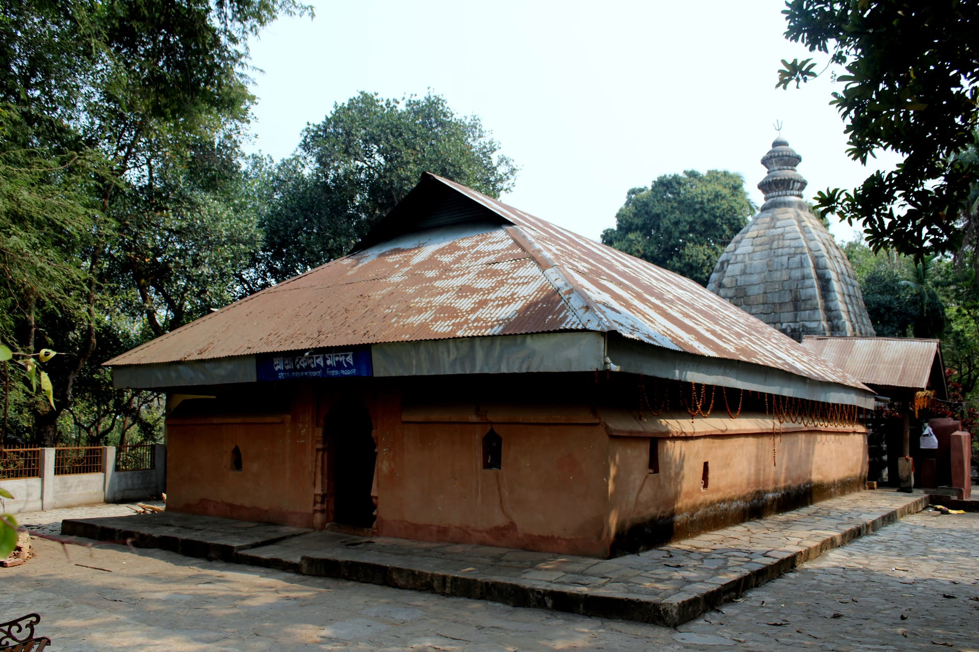 Brick mandapa with a sloped tin roof and, behind it, the stone sikhara of the Kedar temple at Hajo
