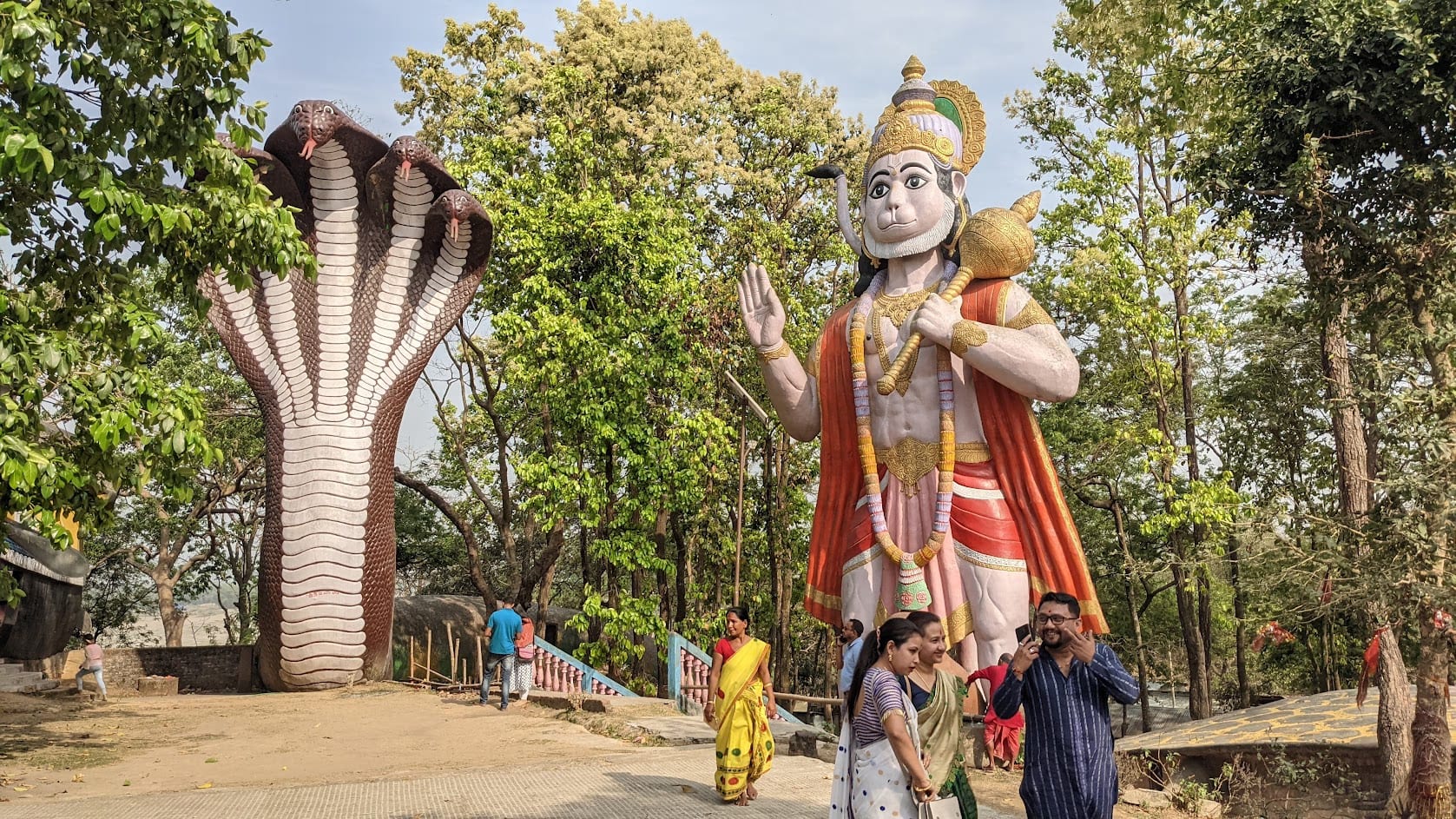 A tall painted Hanuman statue beside a large multi-hooded serpent sculpture among trees, with a few visitors at the base