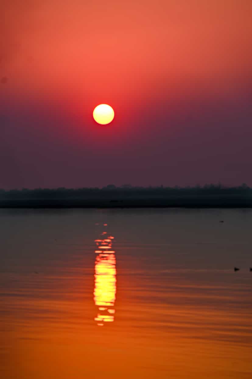 A vivid red-orange sun low over the wide Brahmaputra at Majuli, its glowing reflection running down the still water toward the viewer, the island's low tree-lined bank a dark silhouette across the horizon