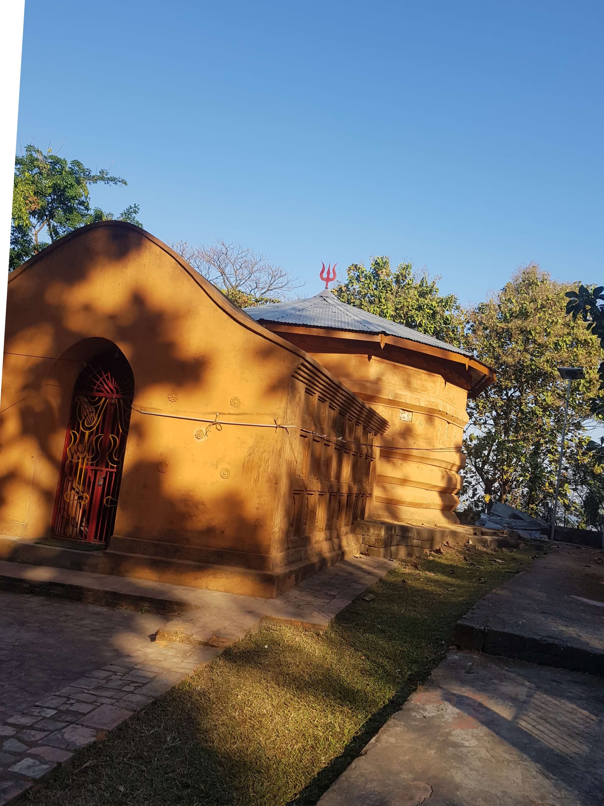 Ochre brick Shiva temple with a curved roof and circular sanctum crowned by a trident, on a grassy hilltop in warm golden light under a clear blue sky