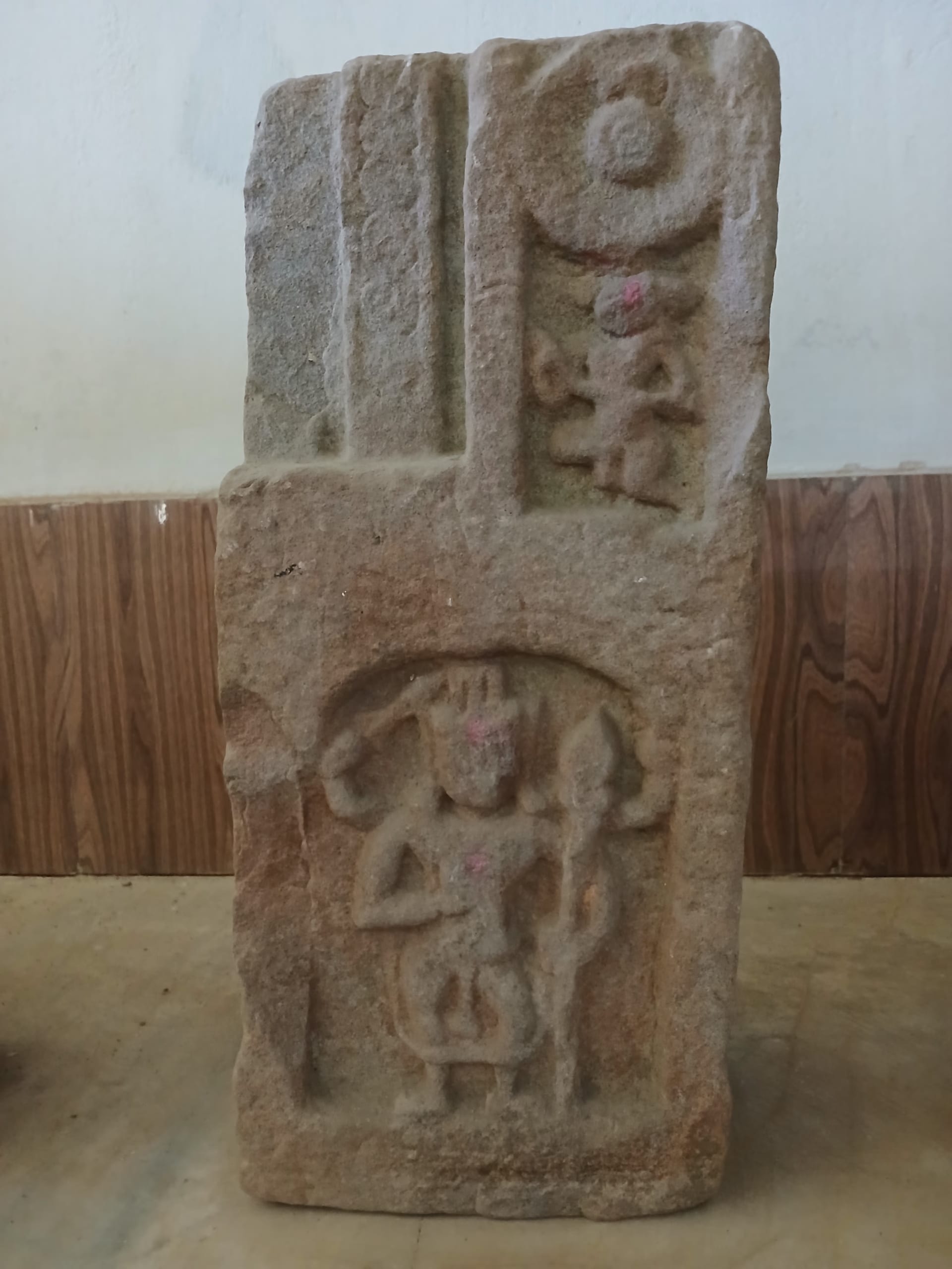 Tall sandstone pillar from the Merghar site standing on a museum floor, its lower face carved with a standing many-armed deity in an arched niche and an upper panel bearing a wheel motif and a smaller figure