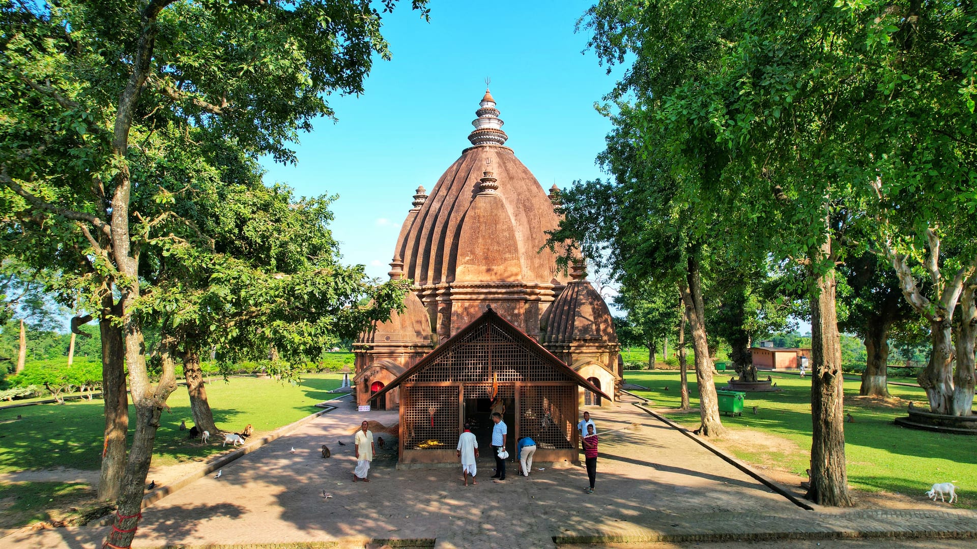 The Negheriting temple seen from a slight rise, its ribbed brick tower and tiered porch framed by green trees under a clear blue sky, with the corner subsidiary shrines flanking the central dol