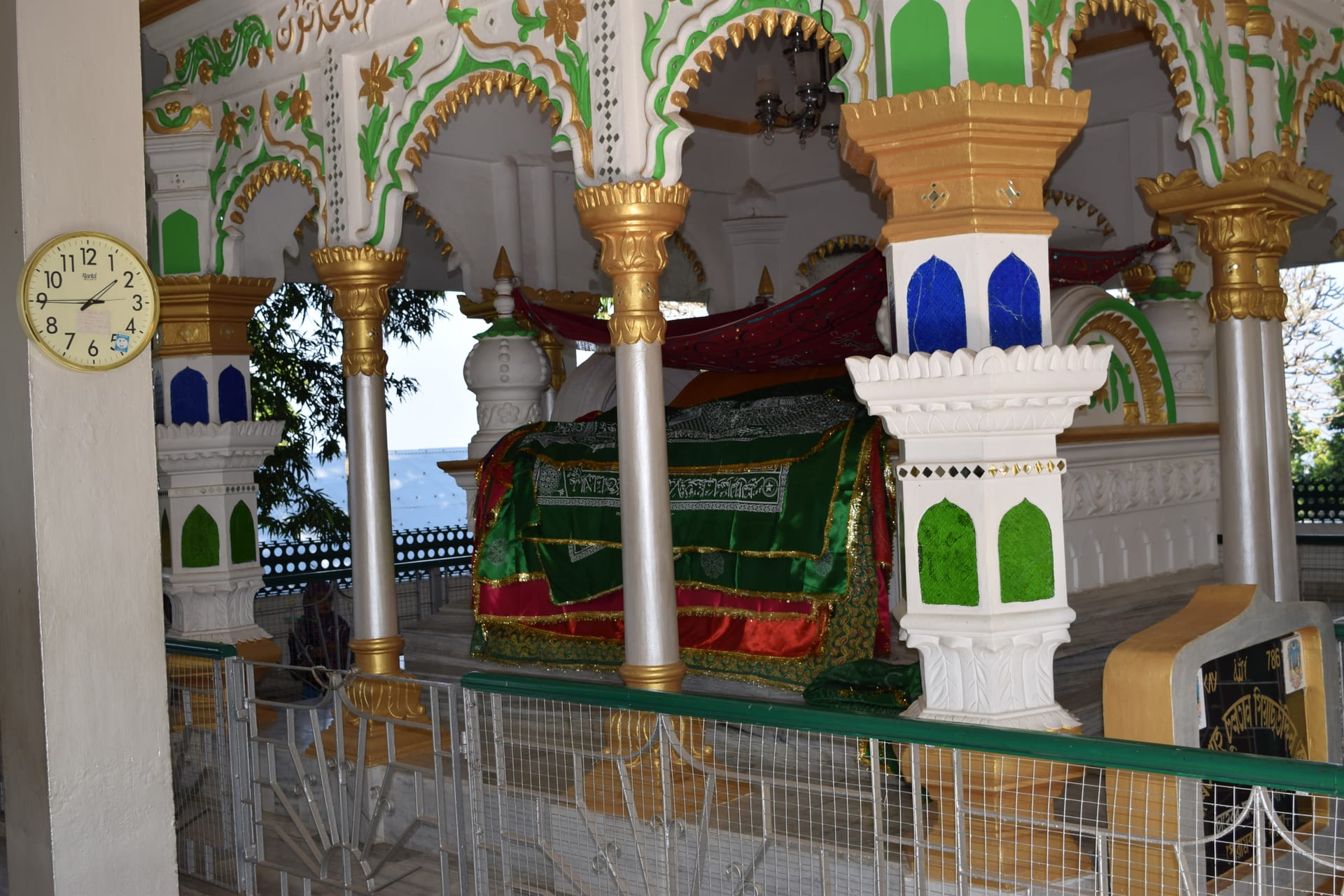 The saint's grave inside the dargah, draped in a green-and-red embroidered chador, beneath ornate gilt-and-green painted arches and marble pillars, with the hills visible beyond