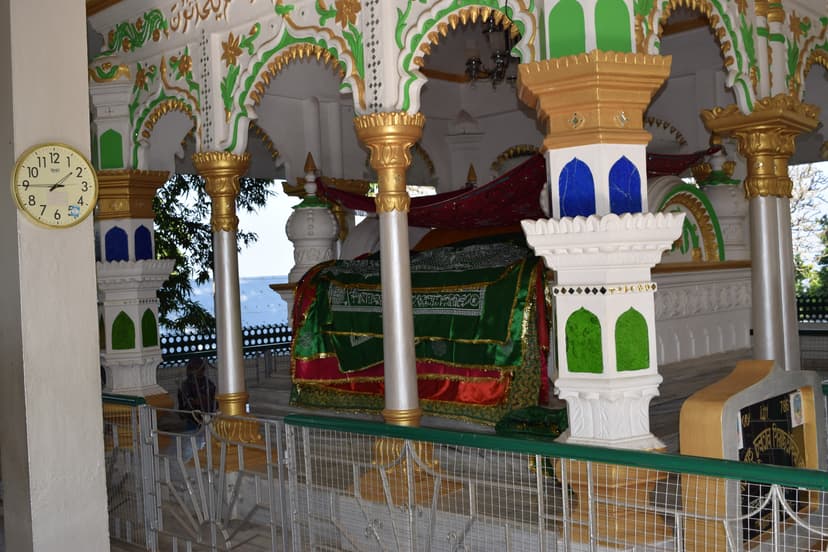 The saint's grave inside the dargah, draped in a green-and-red embroidered chador, beneath ornate gilt-and-green painted arches and marble pillars, with the hills visible beyond
