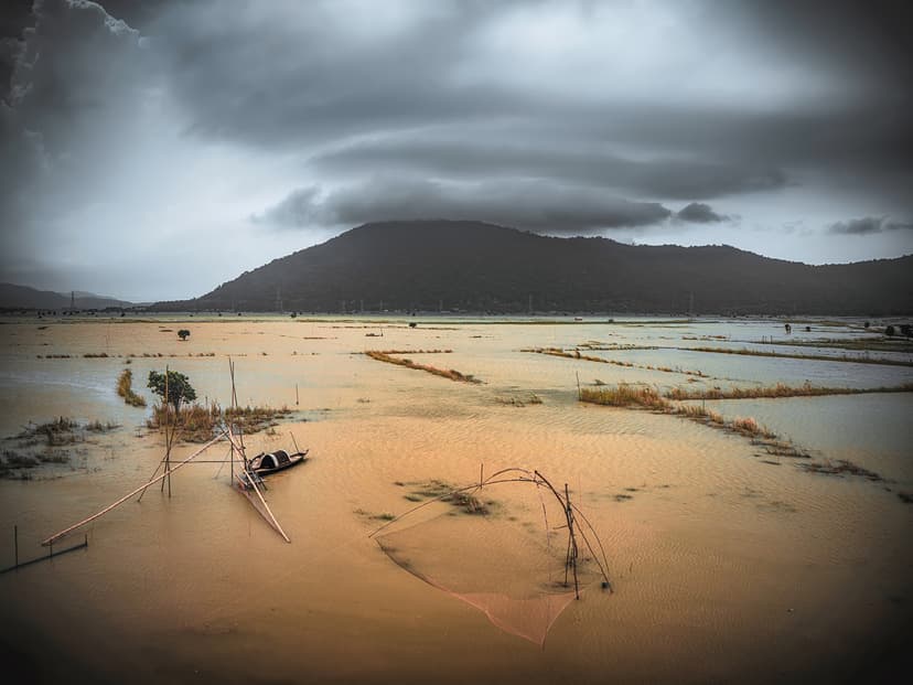 Amber floodwater spreading across Pobitora's grassland below a hill under a heavy monsoon sky, with a small boat and fishing poles