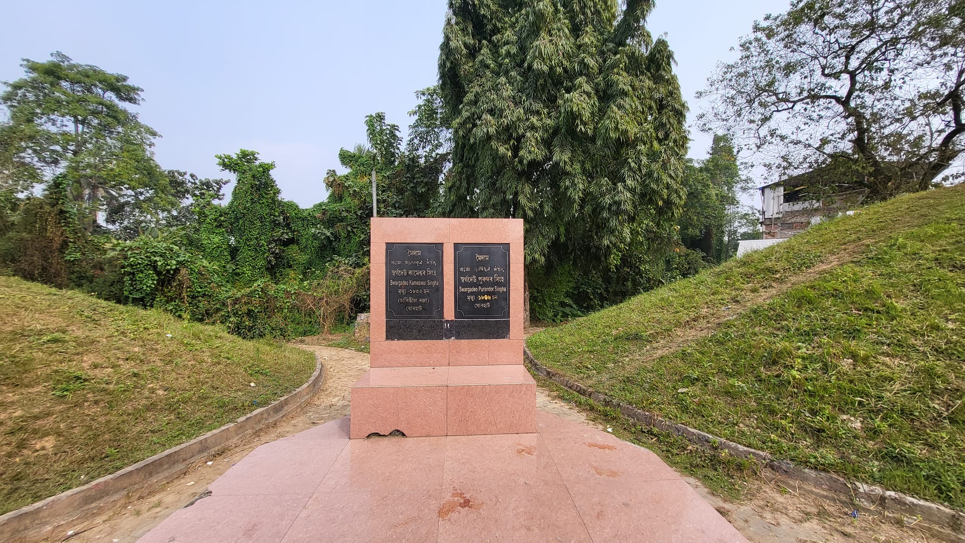 Two grassy earthen Ahom burial mounds flanking a pink-granite plinth with black name-plaques for kings Kameswar Singha and Purandar Singha, Rajamaidam, Jorhat