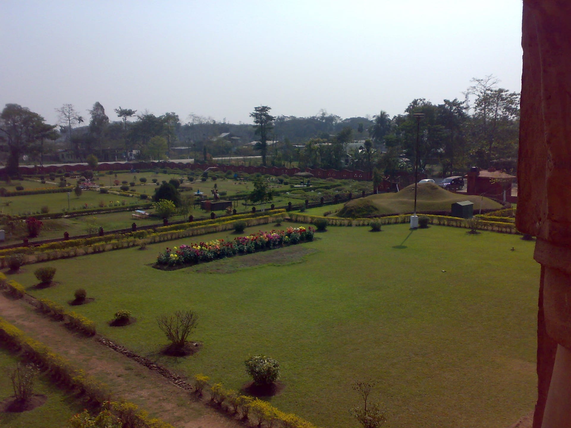 View looking down from the upper gallery of the Rang Ghar over the landscaped gardens, with geometric flower beds, clipped hedges, an open lawn and a tree line beyond