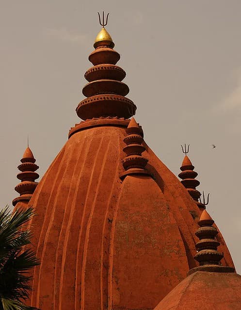 Close view of the red-ochre upper spire of the Sivadol at Sivasagar, crowned by a polished golden finial with stacked disc tiers and smaller corner finials against a pale sky