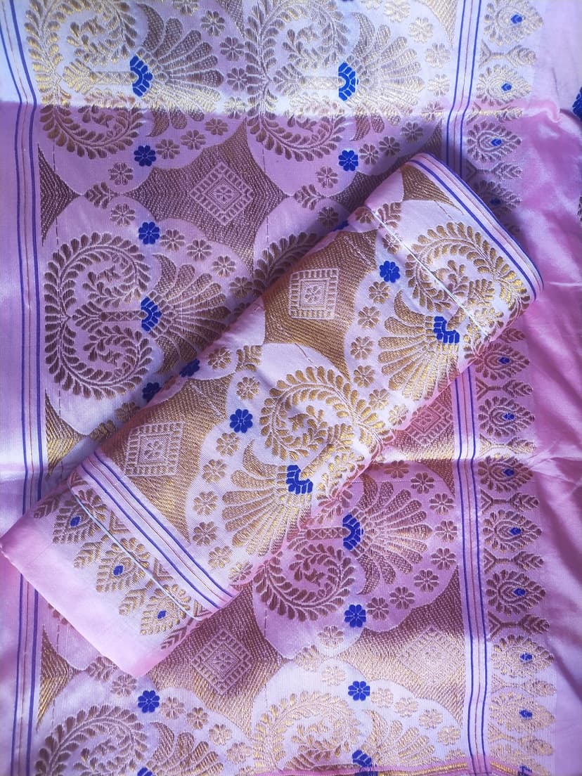 A lavender-pink Assamese mekhela sador with dense gold floral motifs and blue accents, part of the cloth rolled across the field