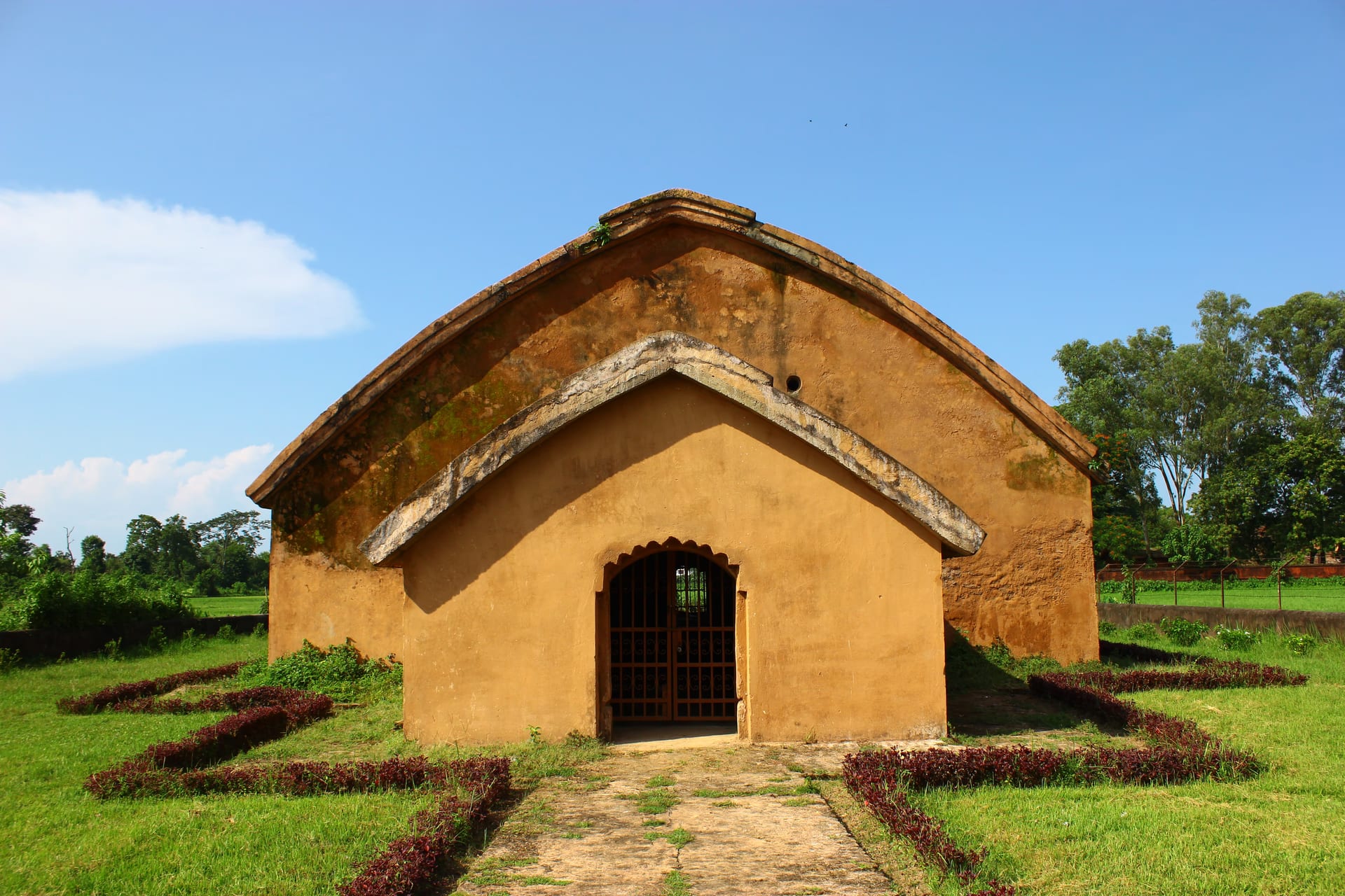 The freestanding do-chala Gola Ghar powder magazine at Rangpur, Sivasagar, ochre plaster walls, curved roof, and gated arched porch on a lawn path