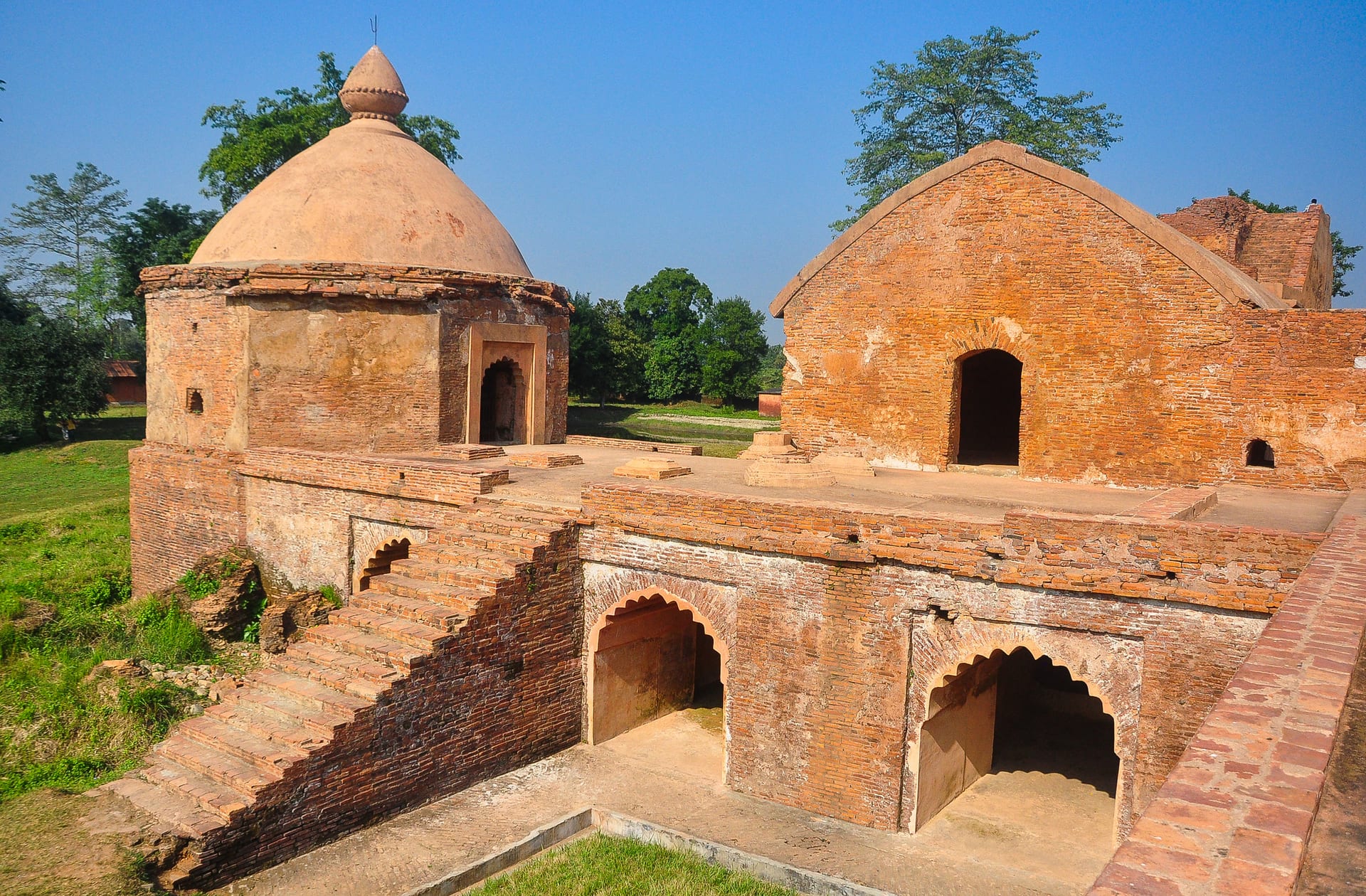 Brick ruins of the Talatal Ghar with a domed pavilion, a descending brick staircase, and cusped arched lower-storey openings under a blue sky