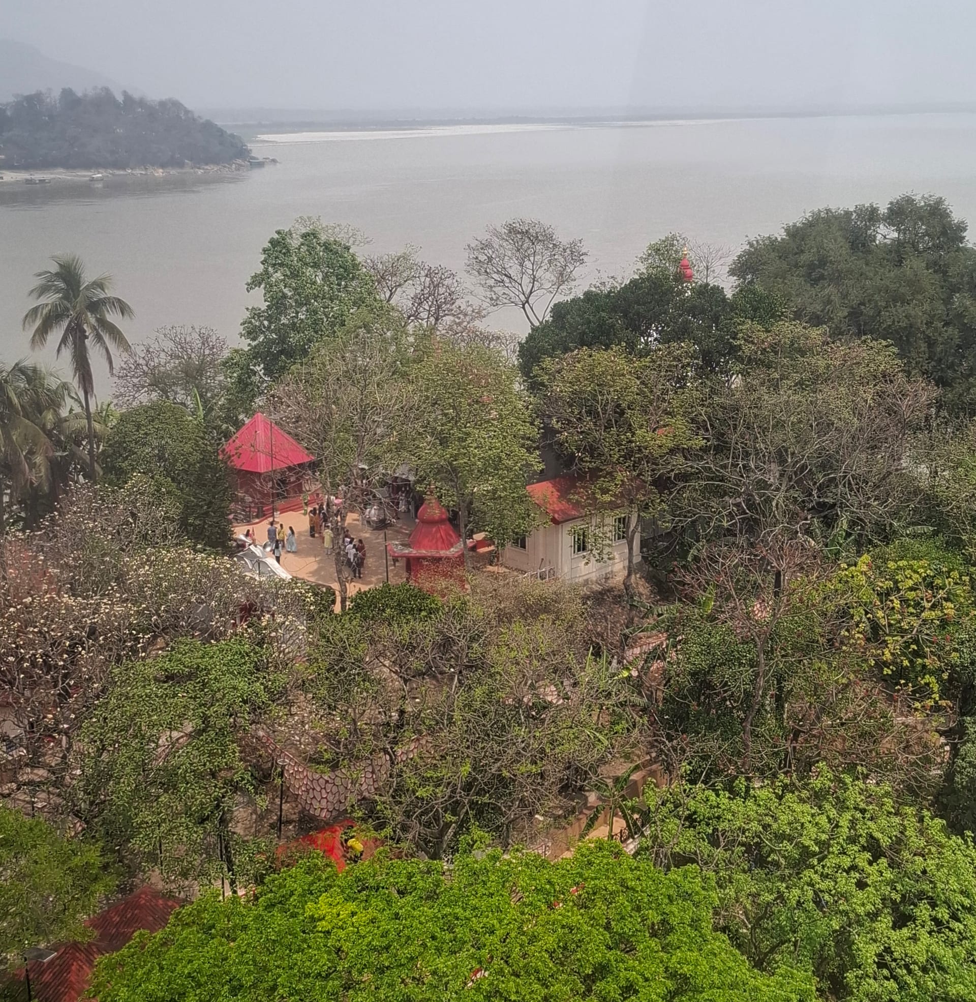 Elevated view of Sukreswar temple's red-roofed shrines on the Brahmaputra bank, with wooded Peacock Island and the Umananda shrine across the water