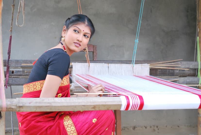 A woman in a traditional Assamese mekhela sador seated at a wooden handloom, a red-and-white gamosa taking shape in the warp before her
