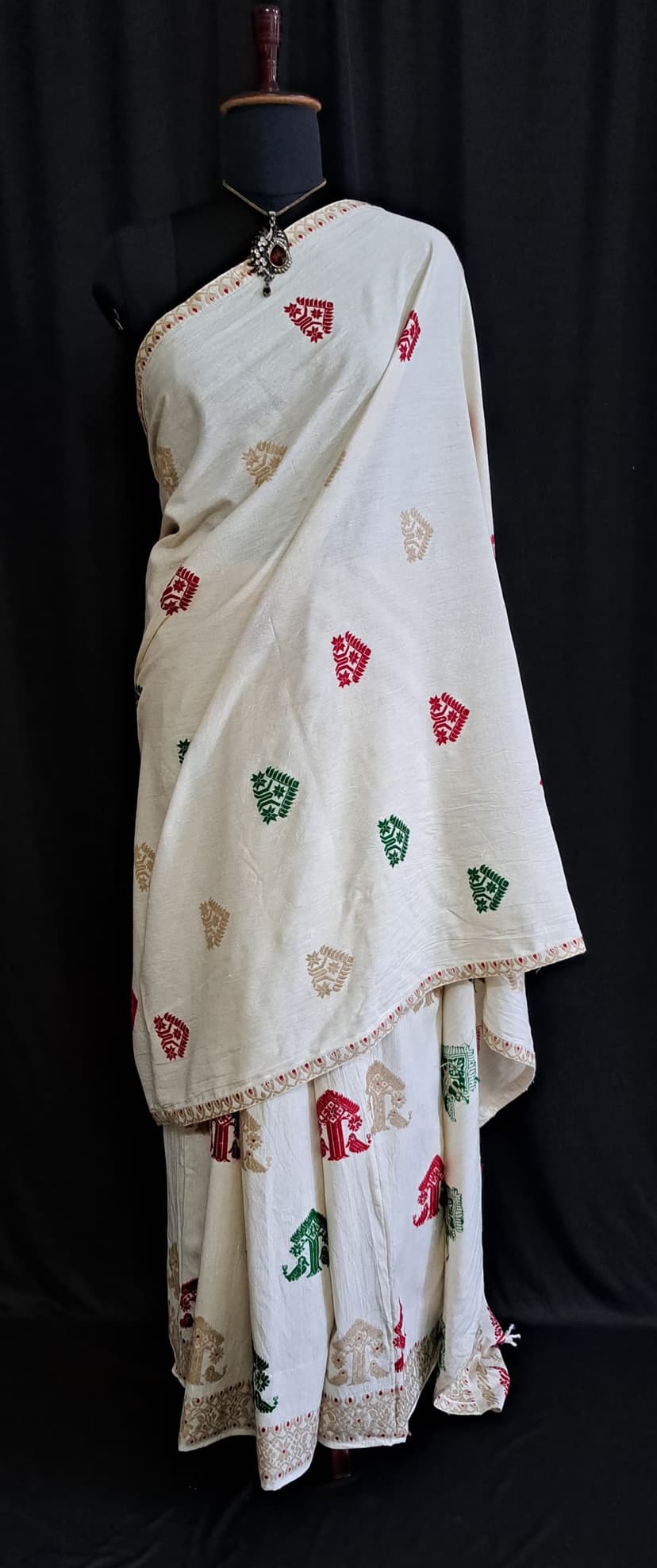 An ivory-white Assamese mekhela sador draped on a stand, its white field scattered with small woven motifs in red, green and gold