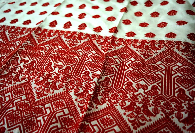 A white pat silk mekhela sador spread flat, its ivory field covered with dense hand-woven red floral and geometric motifs, scattered red butis on the plainer panel above