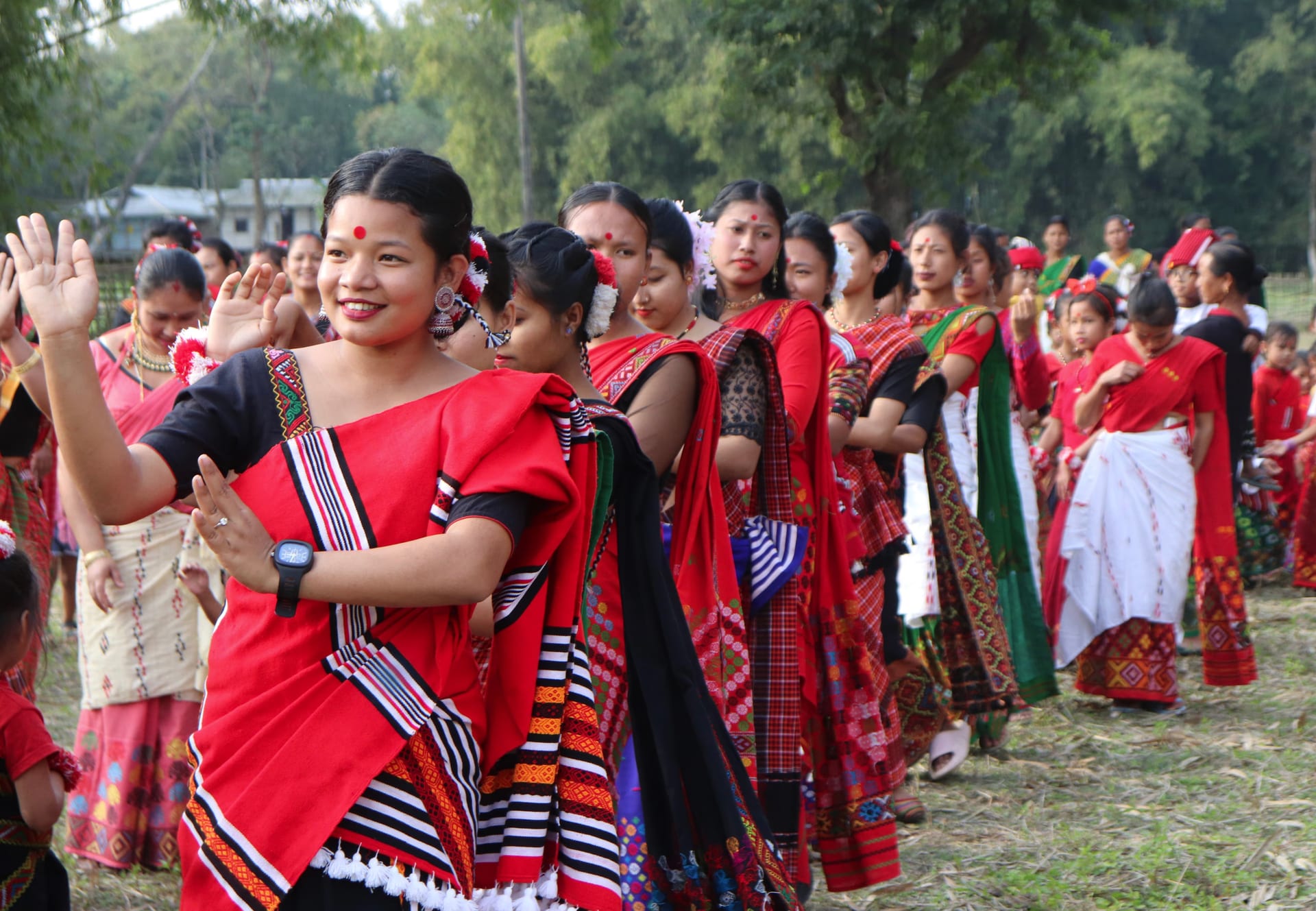 A line of young Mising women in red and striped traditional woven dress dancing in an open field