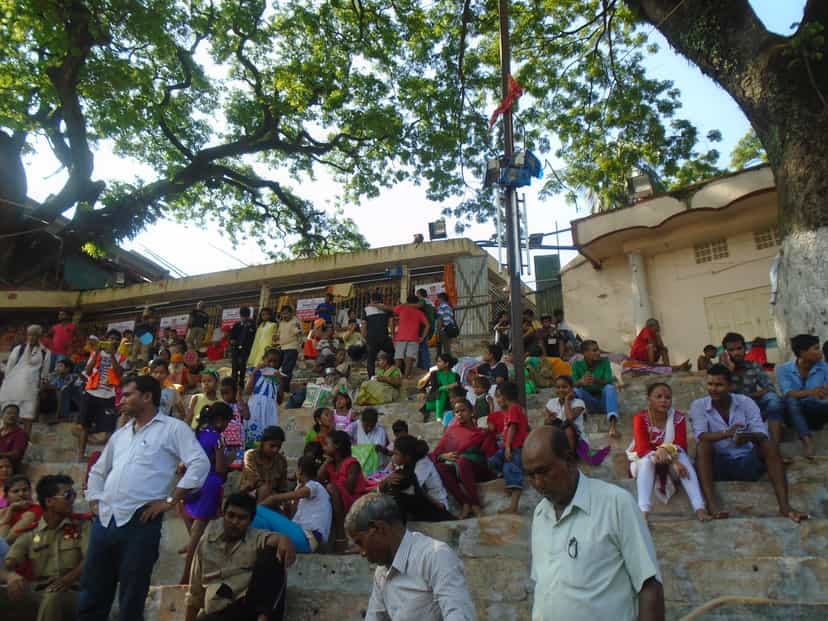 Crowds of pilgrims seated and standing on the broad stone steps beneath a large spreading tree at the Kamakhya complex during the Ambubachi Mela, festival banners and shopfronts behind