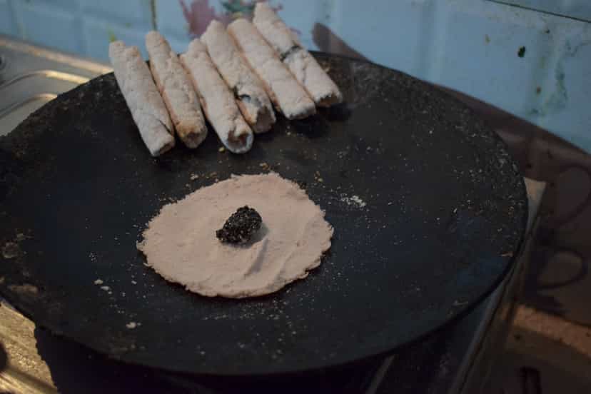 Rolled til pitha rice-cakes resting on a black griddle, with a thin rice batter crepe and a mound of black sesame filling being formed