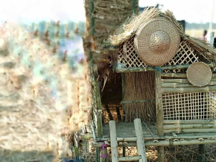 A makeshift hut of straw, thatch and bamboo raised in a harvested field, with woven bamboo trays and a conical hat hung on its frame
