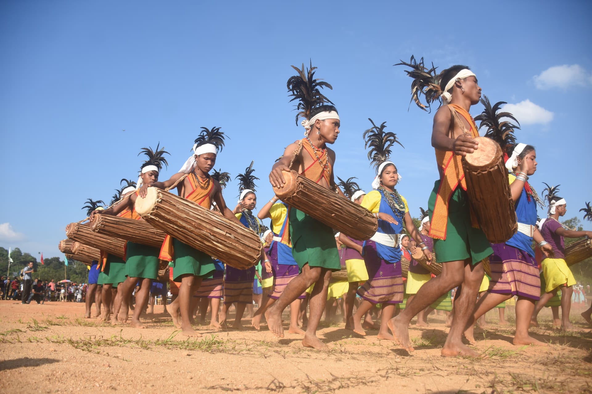 Lines of Garo dancers in feathered headdresses, the men carrying long oval dama drums, moving in formation across an open field
