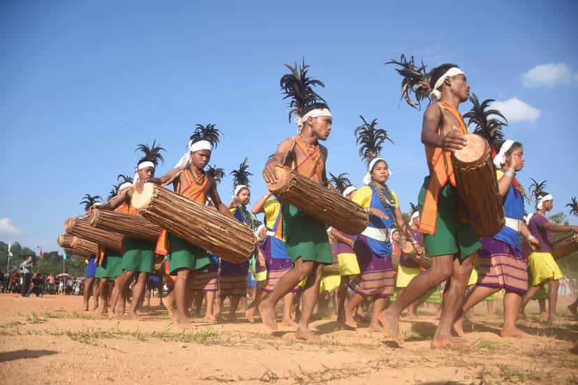 Lines of Garo dancers in feathered headdresses, the men carrying long oval dama drums, moving in formation across an open field