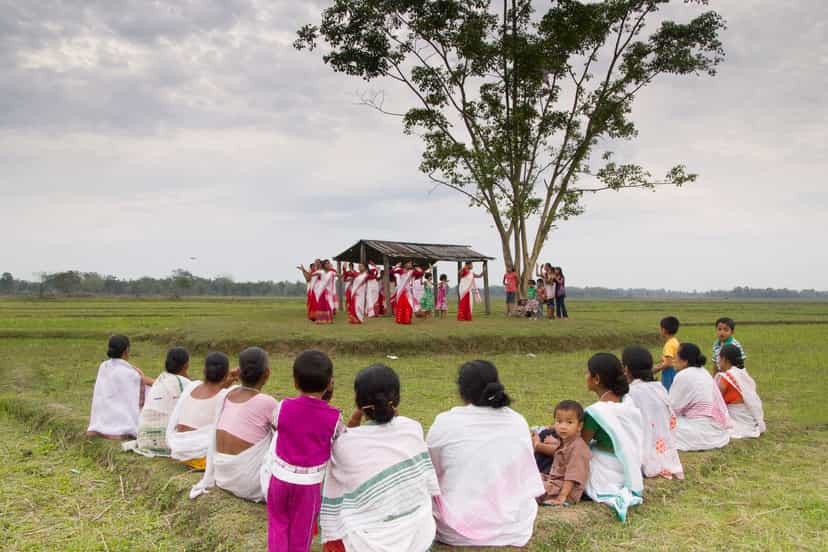 Women in red-and-white mekhela-chador dancing under a tree beside a thatched field shelter in an open green paddy field, with rows of villagers wrapped in white gamosa seated on the grass watching from the foreground under a grey sky